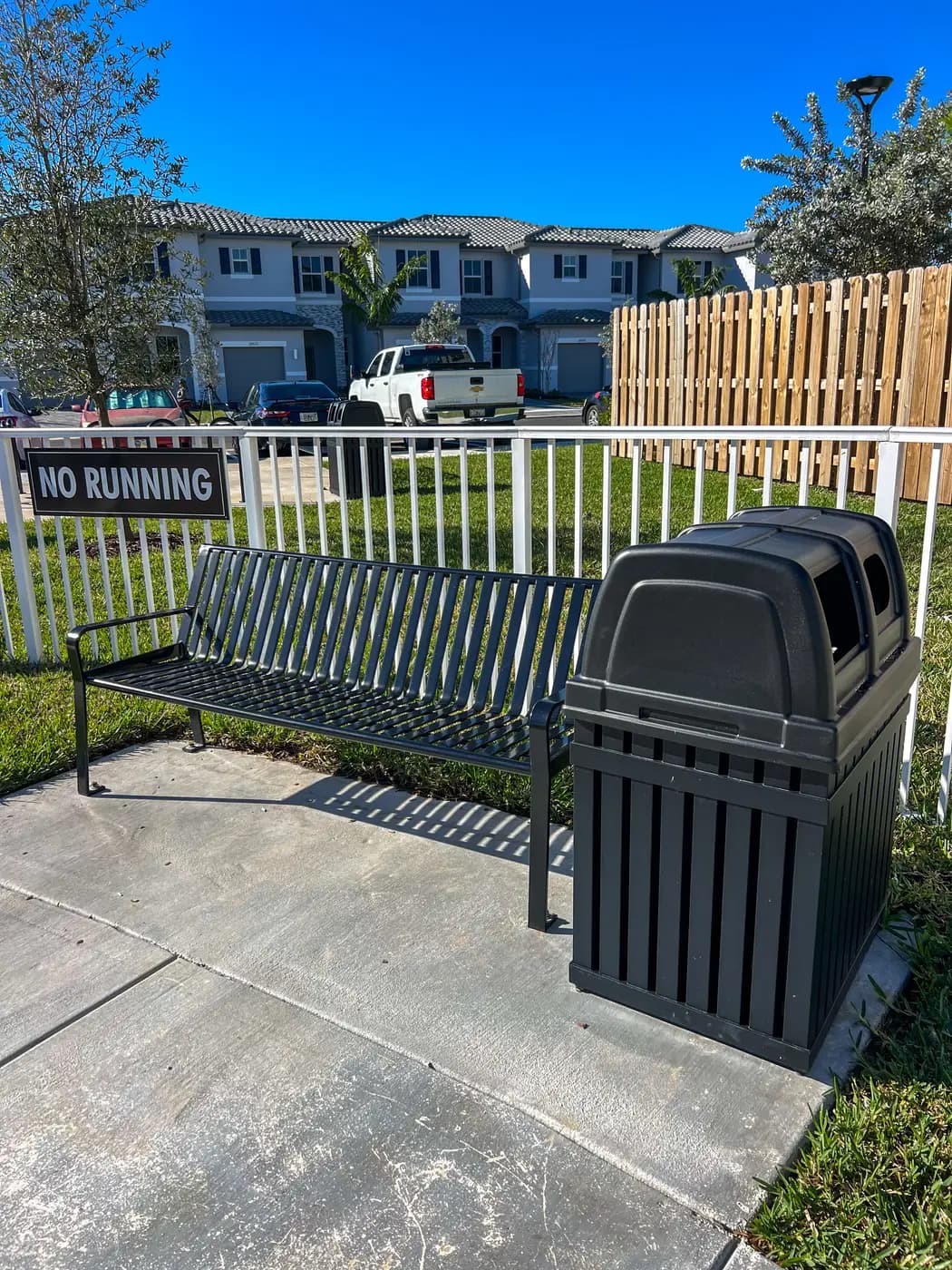 Metal bench and covered trash receptacle beside a community playground fence
